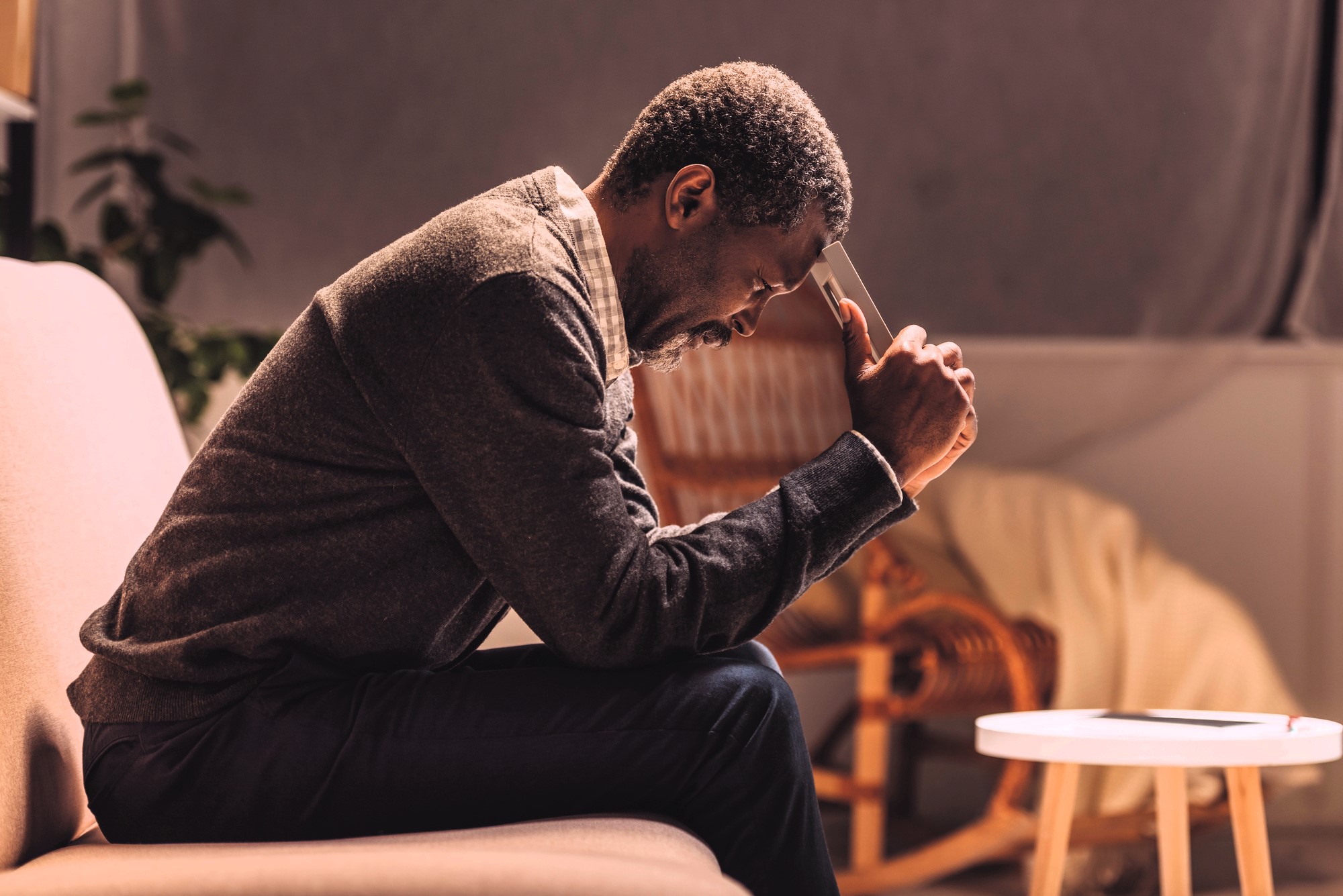 A man sitting on a sofa, leaning forward with his elbows on his knees and holding a smartphone to his forehead. He appears deep in thought or concerned. The room is softly lit with neutral-toned furnishings in the background.