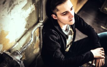 Young man in a black hoodie sits on the floor against a worn, textured wall, looking contemplative with his head slightly tilted up. The lighting is dim, and he appears thoughtful or introspective.