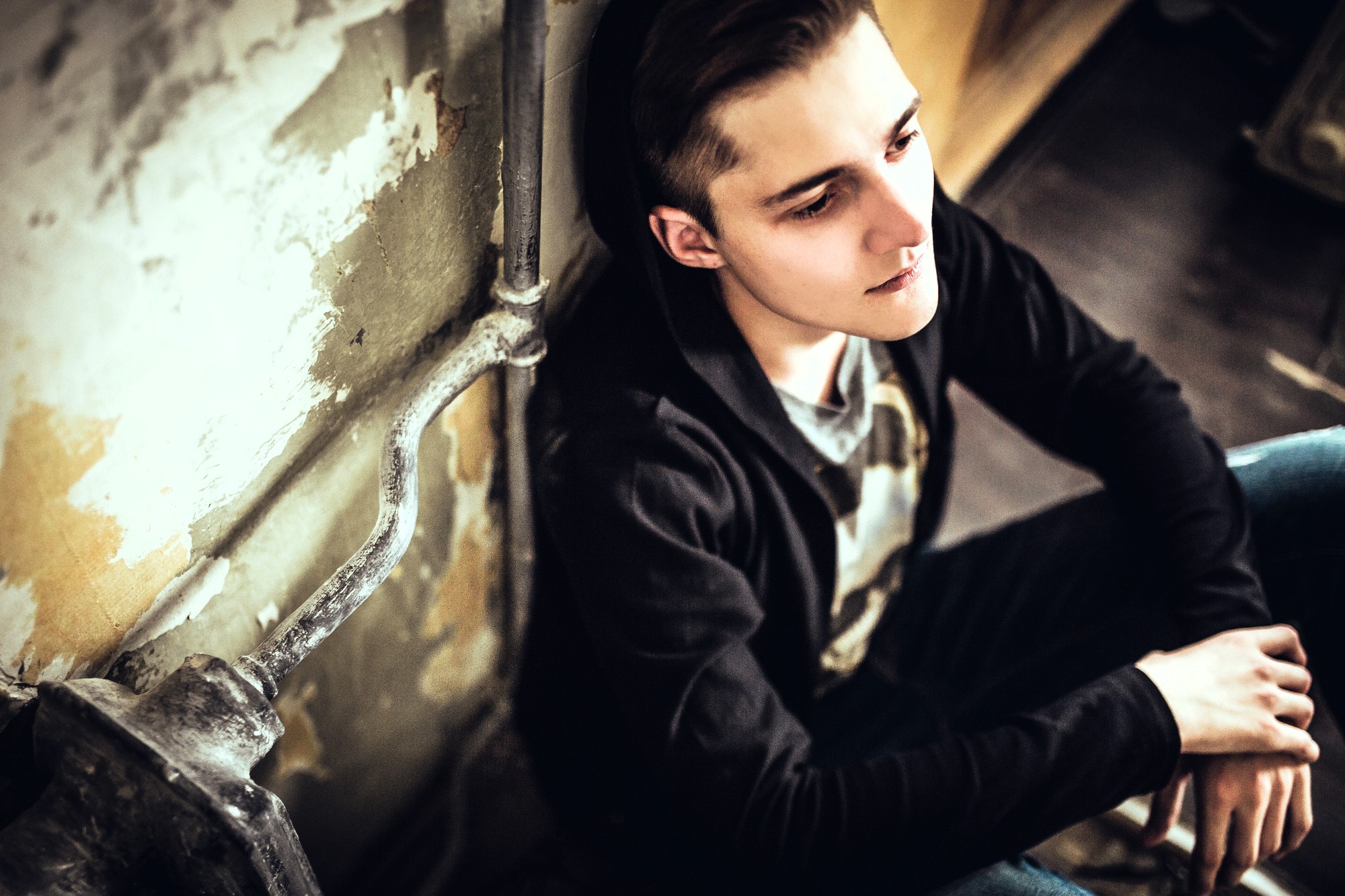 Young man in a black hoodie sits on the floor against a worn, textured wall, looking contemplative with his head slightly tilted up. The lighting is dim, and he appears thoughtful or introspective.