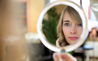 A person with blonde hair is looking into a circular mirror, capturing their reflection. They appear focused, with soft lighting. The background is blurred with hints of indoor plants and furnishings.