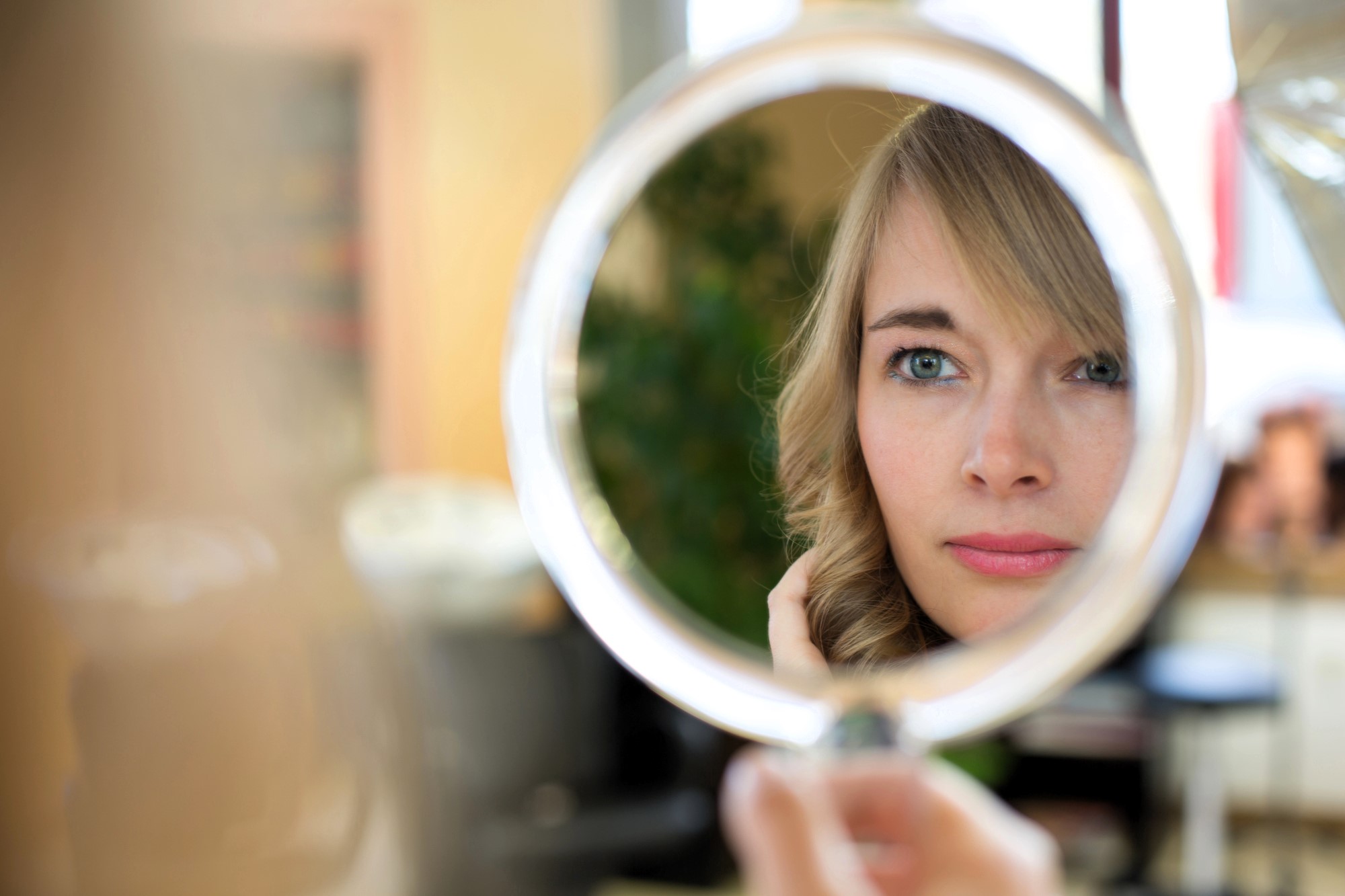 A person with blonde hair is looking into a circular mirror, capturing their reflection. They appear focused, with soft lighting. The background is blurred with hints of indoor plants and furnishings.