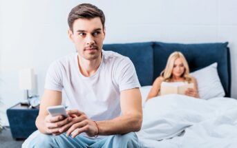 A man sits on a bed holding a smartphone, looking away thoughtfully. Behind him, a woman sits up in bed reading a book. The room is modern and well-lit, with a blue headboard and white bedding.