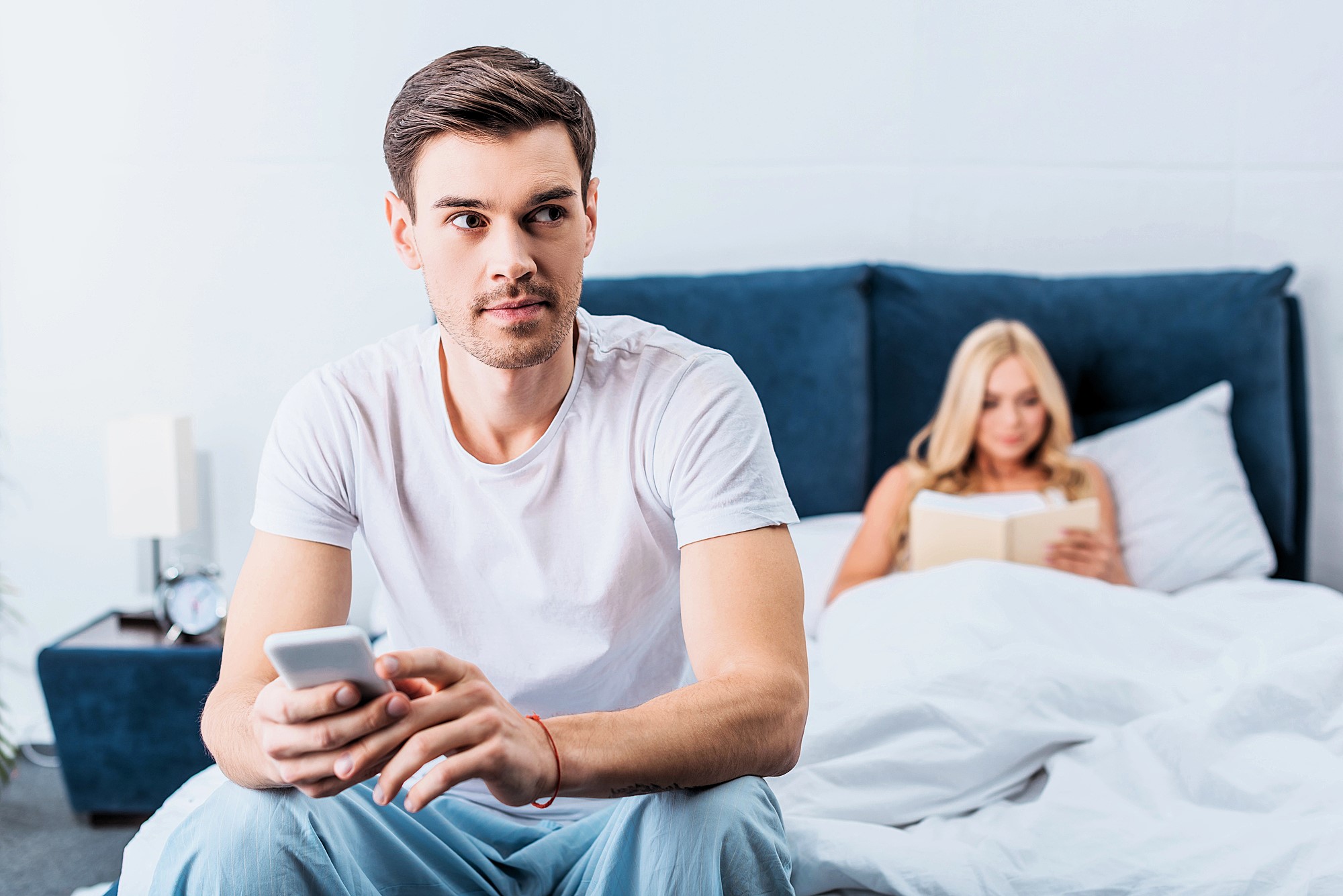 A man sits on a bed holding a smartphone, looking away thoughtfully. Behind him, a woman sits up in bed reading a book. The room is modern and well-lit, with a blue headboard and white bedding.