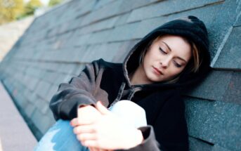 A person wearing a black hoodie is sitting against a concrete wall, looking down with a contemplative expression. The background features a slanted, textured surface, and there is a soft, outdoor light.