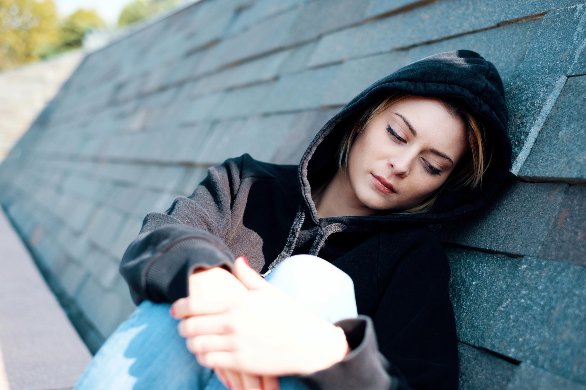 A person wearing a black hoodie is sitting against a concrete wall, looking down with a contemplative expression. The background features a slanted, textured surface, and there is a soft, outdoor light.