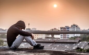 A person in a hooded sweatshirt and jeans sits on a rooftop, resting their arms on their knees. The sun is low in the sky, casting a warm glow over the buildings in the background.