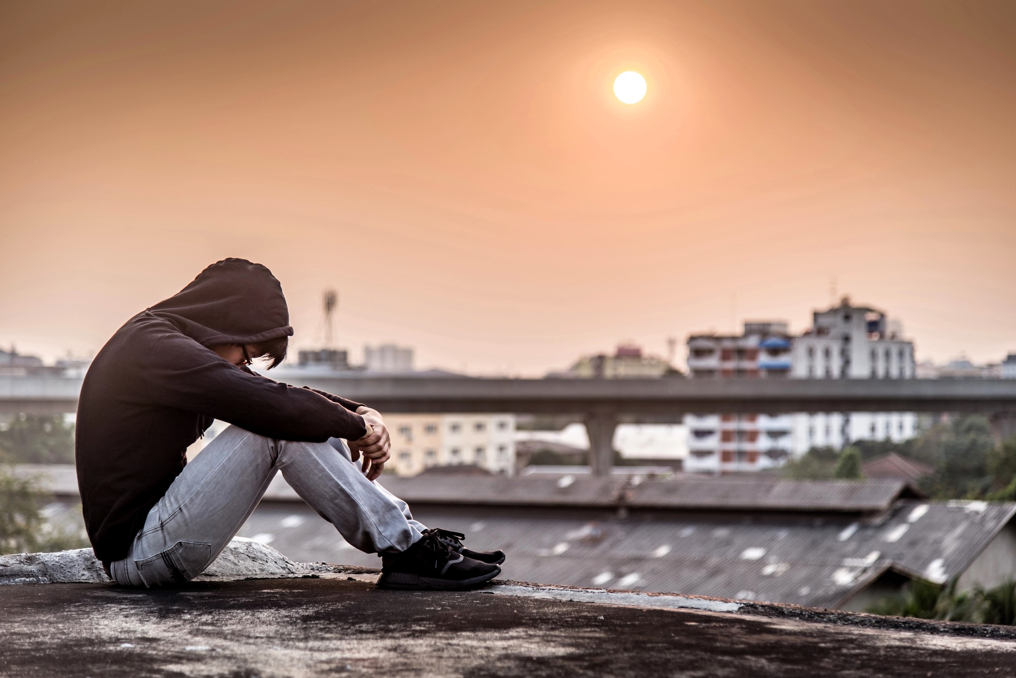 A person in a hooded sweatshirt and jeans sits on a rooftop, resting their arms on their knees. The sun is low in the sky, casting a warm glow over the buildings in the background.