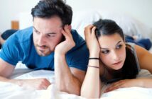 A man and a woman lie on a bed, both looking thoughtful or concerned. The man rests his head on his hand, while the woman holds her head with one hand. They are both wearing casual clothes, and the background is simple and neutral.