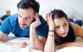 A man and a woman lie on a bed, both looking thoughtful or concerned. The man rests his head on his hand, while the woman holds her head with one hand. They are both wearing casual clothes, and the background is simple and neutral.