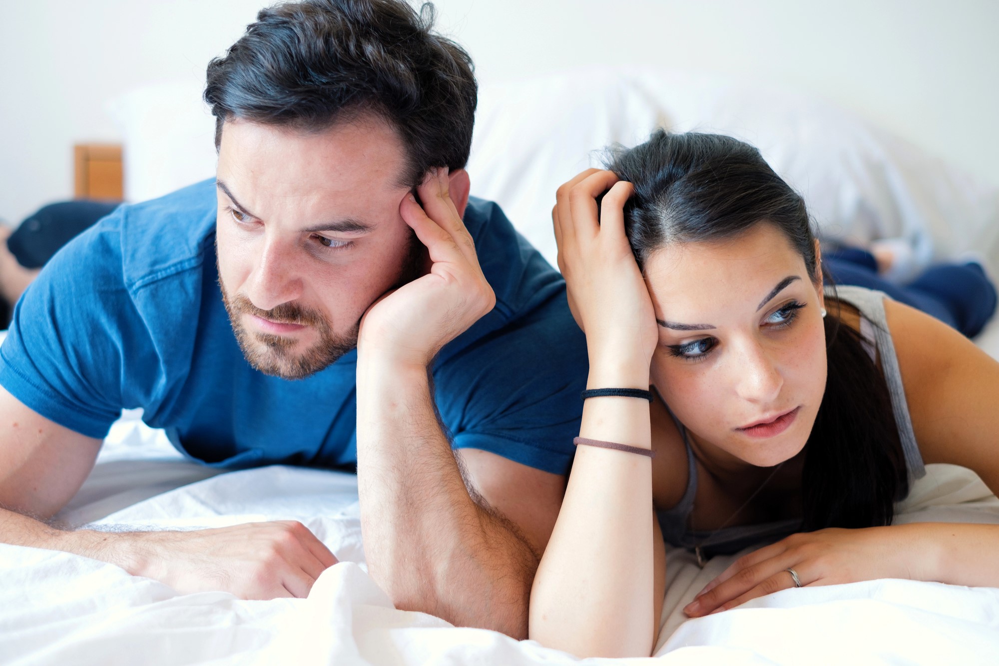 A man and a woman lie on a bed, both looking thoughtful or concerned. The man rests his head on his hand, while the woman holds her head with one hand. They are both wearing casual clothes, and the background is simple and neutral.