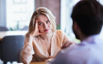 A woman with blonde hair is sitting at a table, resting her head on her hand, looking bored or frustrated. She is wearing a light peach blouse and is facing a man whose back is to the camera. They're in a bright indoor setting.