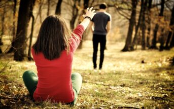 A person sits on the grass with their back to the camera, reaching out toward another person walking away through the forest. The setting is filled with trees and dappled sunlight, creating a warm, introspective atmosphere.