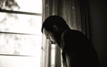 Silhouette of a person with a beard, leaning forward near a window, captured in black and white. The scene is indoors, with soft light filtering through sheer curtains. The mood appears contemplative.