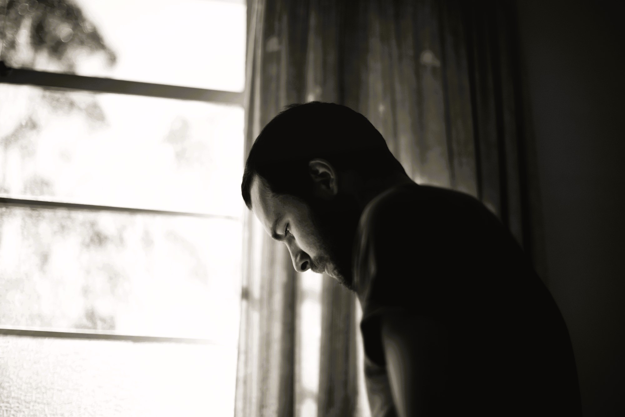 Silhouette of a person with a beard, leaning forward near a window, captured in black and white. The scene is indoors, with soft light filtering through sheer curtains. The mood appears contemplative.