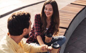 A man and a woman are sitting on a curved wooden bench, smiling and holding paper coffee cups. The woman is wearing a red plaid shirt and jeans, while the man is in a yellow shirt. They appear to be engaged in a conversation outdoors.