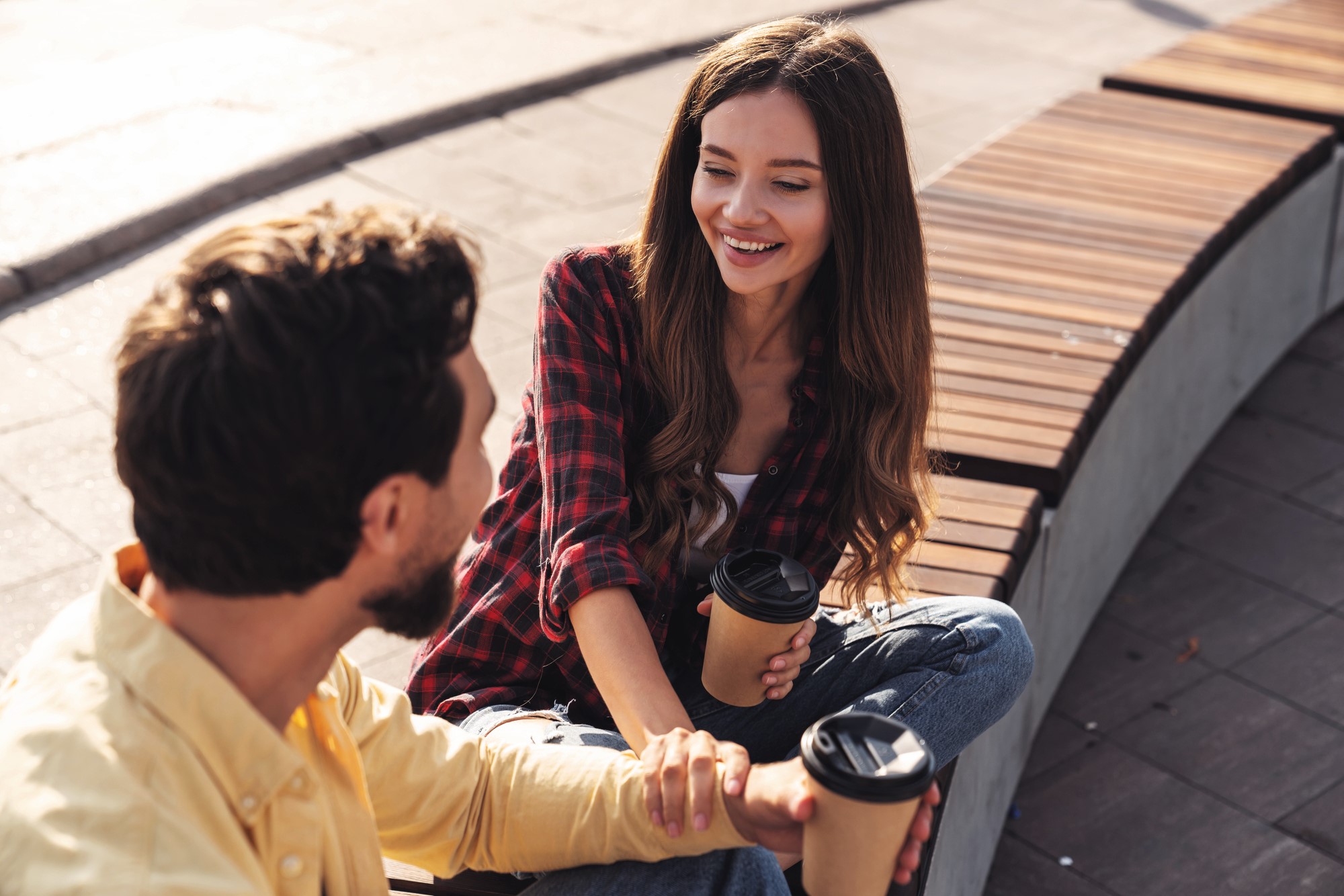 A man and a woman are sitting on a curved wooden bench, smiling and holding paper coffee cups. The woman is wearing a red plaid shirt and jeans, while the man is in a yellow shirt. They appear to be engaged in a conversation outdoors.