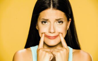 A woman with long dark hair wearing a light blue tank top is pressing her fingers into her cheeks, forcing a smile. She looks slightly uncomfortable. The background is a solid yellow color.