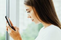 A young woman with long brown hair holds a smartphone and looks intently at the screen. She is standing by a window, with a soft focus view of greenery outside. She wears a white top, and the lighting is bright and natural.