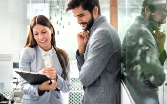 A woman and a man in business attire stand in an office. The woman smiles and holds a clipboard, while the man rests his chin on his hand, looking at the clipboard. They are reflected in the glass wall beside them.