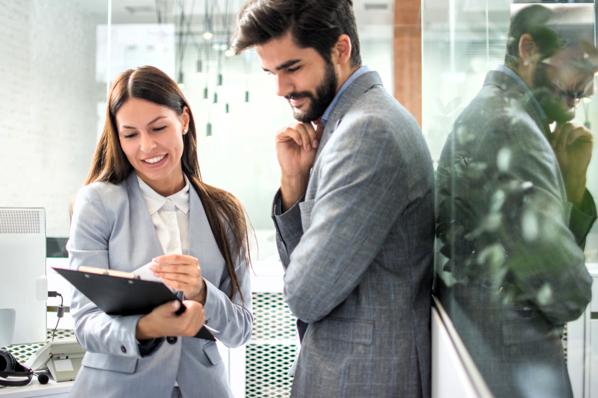 A woman and a man in business attire stand in an office. The woman smiles and holds a clipboard, while the man rests his chin on his hand, looking at the clipboard. They are reflected in the glass wall beside them.