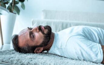 Man lying on a carpeted floor, wearing a white T-shirt, with a contemplative expression. A potted plant is in the blurred background.