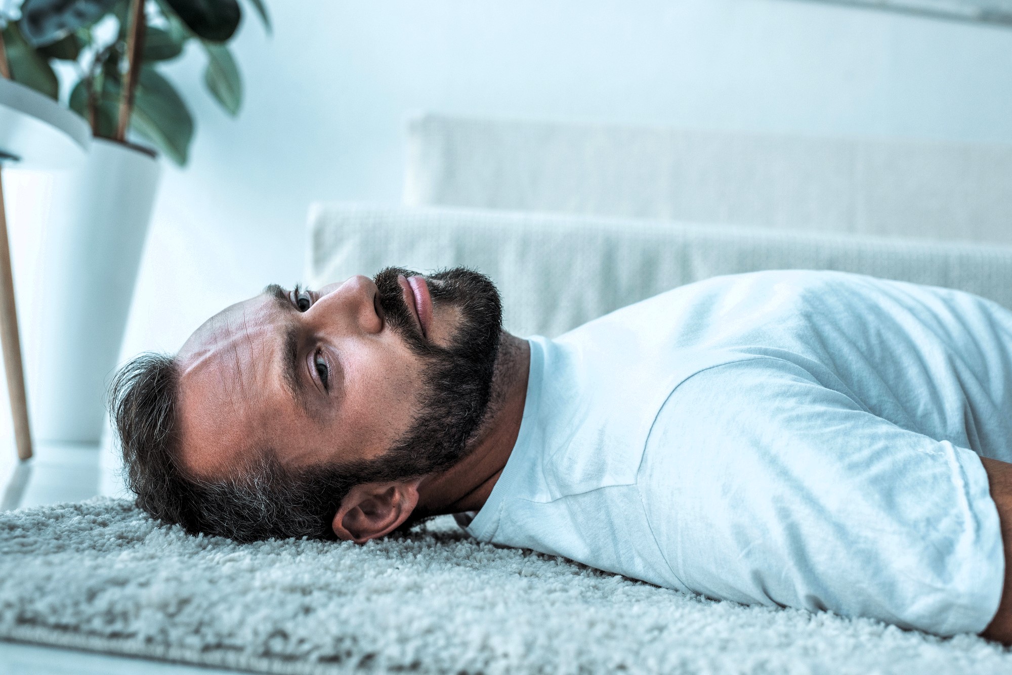 Man lying on a carpeted floor, wearing a white T-shirt, with a contemplative expression. A potted plant is in the blurred background.