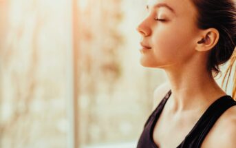 Woman in a black tank top practicing mindfulness with eyes closed against a softly lit window background, suggesting a calm and peaceful setting.