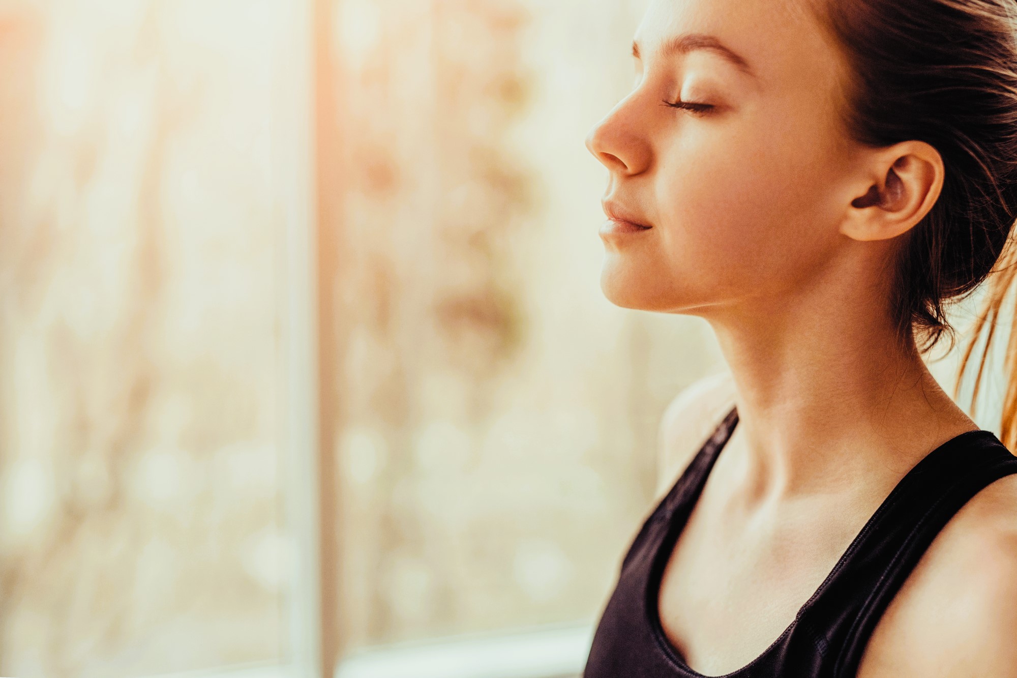 Woman in a black tank top practicing mindfulness with eyes closed against a softly lit window background, suggesting a calm and peaceful setting.