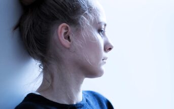 A person with light hair is gazing contemplatively to the right. They are standing against a light-colored background, wearing a dark top. The lighting is soft and highlights the profile of their face.