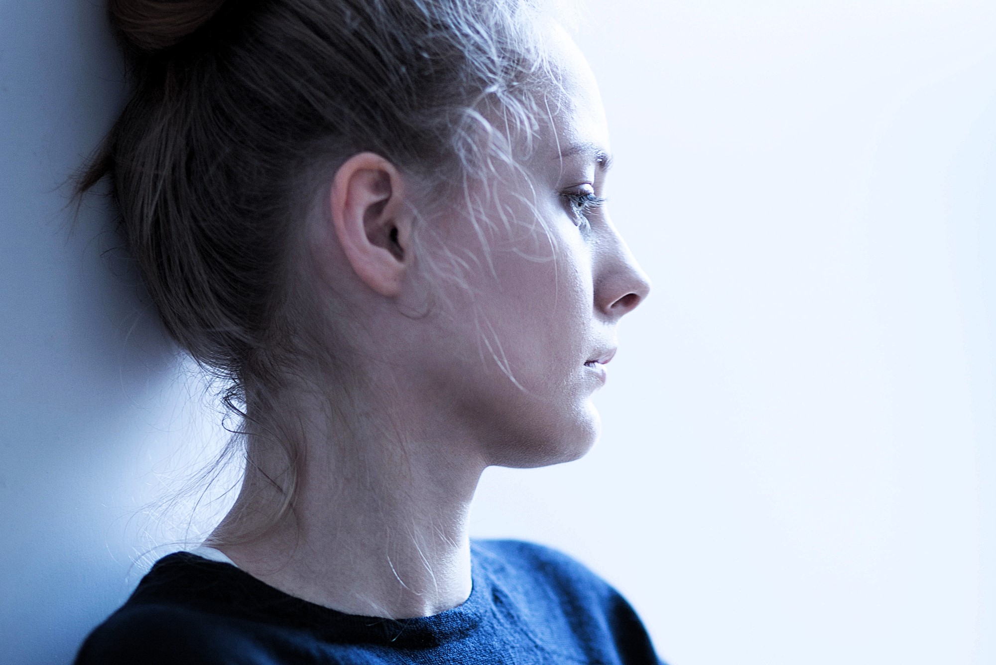 A person with light hair is gazing contemplatively to the right. They are standing against a light-colored background, wearing a dark top. The lighting is soft and highlights the profile of their face.