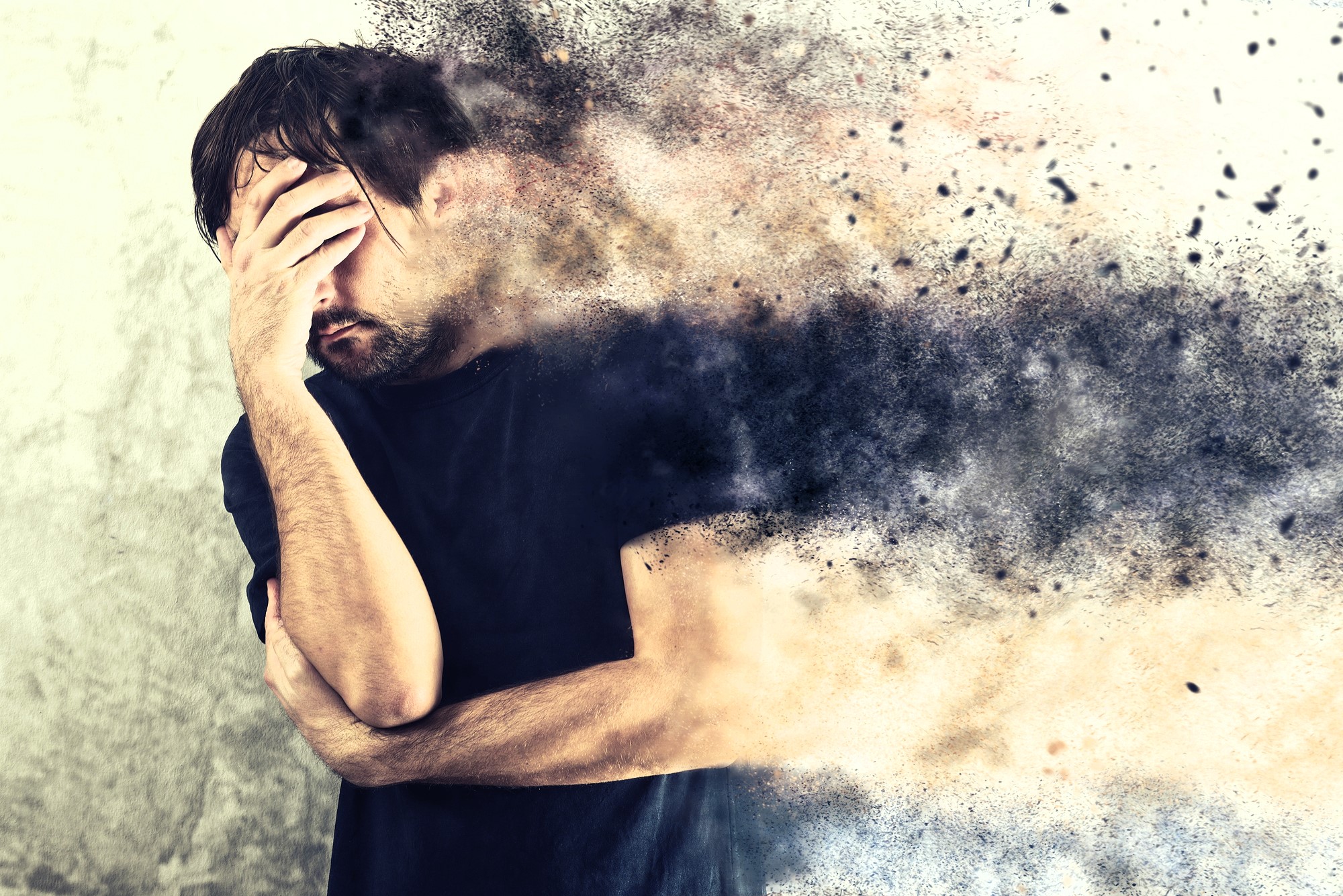 A man holding his head with one hand stands against a textured background. His body appears to dissolve into smoke and particles, creating a dynamic and surreal effect.