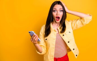 A surprised woman with long brown hair holds a smartphone in one hand and touches her head with the other. She wears a yellow jacket over a striped shirt and red pants, standing against a bright yellow background. Her mouth is open in shock.