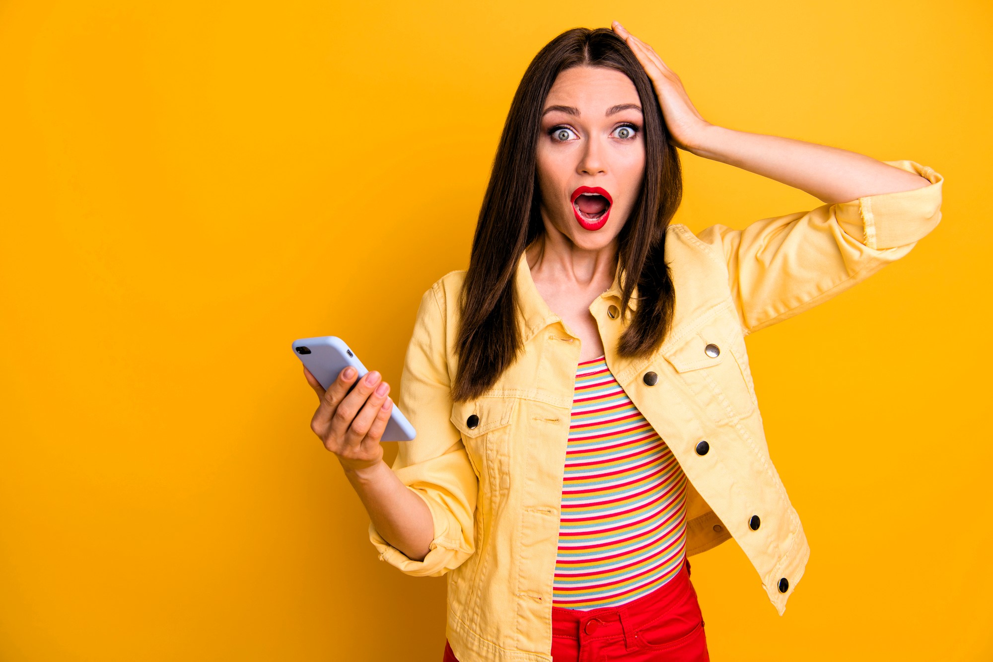 A surprised woman with long brown hair holds a smartphone in one hand and touches her head with the other. She wears a yellow jacket over a striped shirt and red pants, standing against a bright yellow background. Her mouth is open in shock.