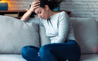 Woman sitting on a couch holding her head in one hand, looking down with a pained expression. She is wearing a striped long-sleeve shirt and jeans, with a plant and blue lamp in the background.