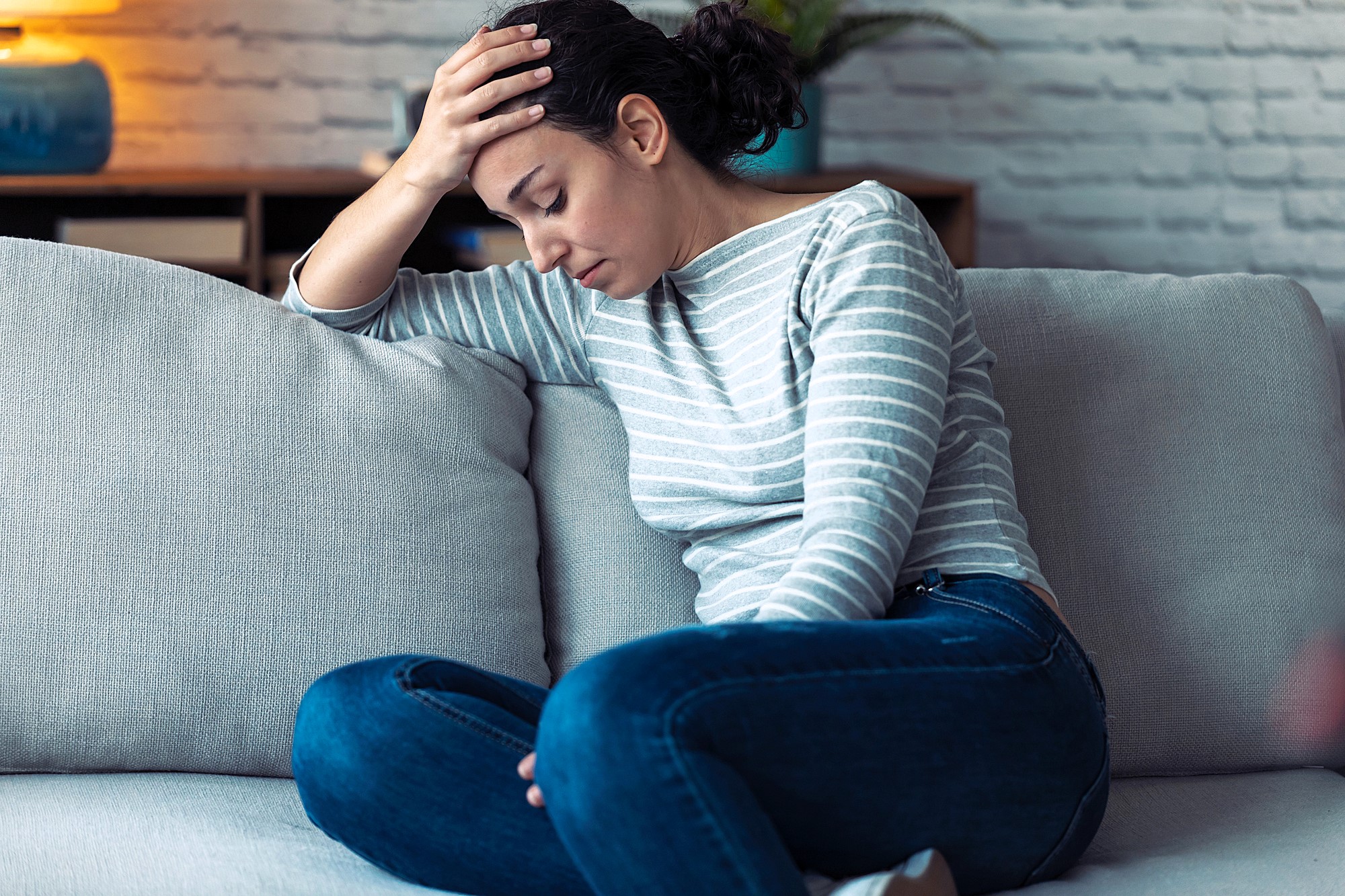 Woman sitting on a couch holding her head in one hand, looking down with a pained expression. She is wearing a striped long-sleeve shirt and jeans, with a plant and blue lamp in the background.