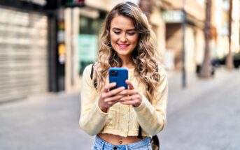 A smiling woman with long, wavy blonde hair looks at her smartphone while standing on a street. She is wearing a light yellow sweater and jeans. The background shows a blurred view of shops and buildings.