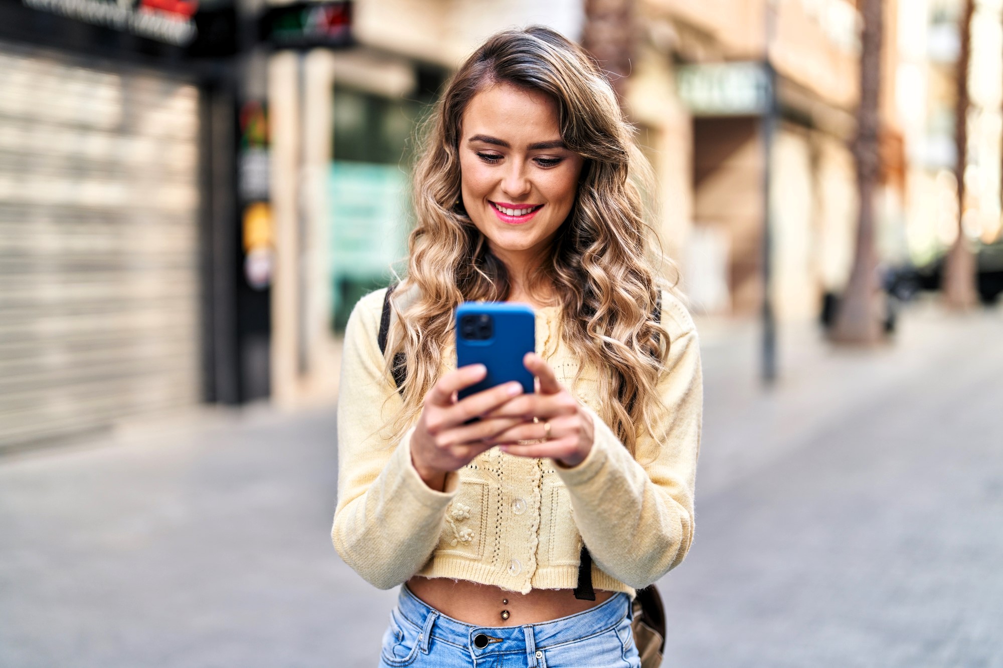 A smiling woman with long, wavy blonde hair looks at her smartphone while standing on a street. She is wearing a light yellow sweater and jeans. The background shows a blurred view of shops and buildings.