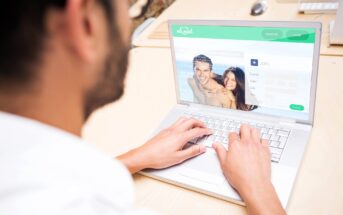 A person is using a laptop at a desk, looking at a dating website. The screen shows a sign-up page with a smiling couple's image and fields for name, date of birth, and city. The desk has a partial view of other devices.