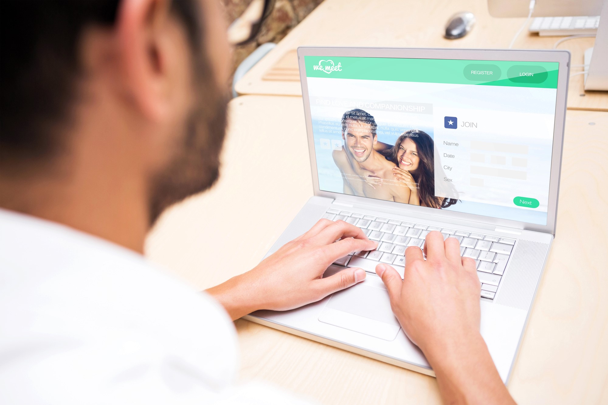 A person is using a laptop at a desk, looking at a dating website. The screen shows a sign-up page with a smiling couple's image and fields for name, date of birth, and city. The desk has a partial view of other devices.