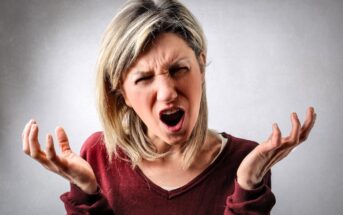 A frustrated woman with blond hair wearing a maroon top stands against a gray background. She has an open mouth and furrowed brows, raising both hands in an expressive gesture.