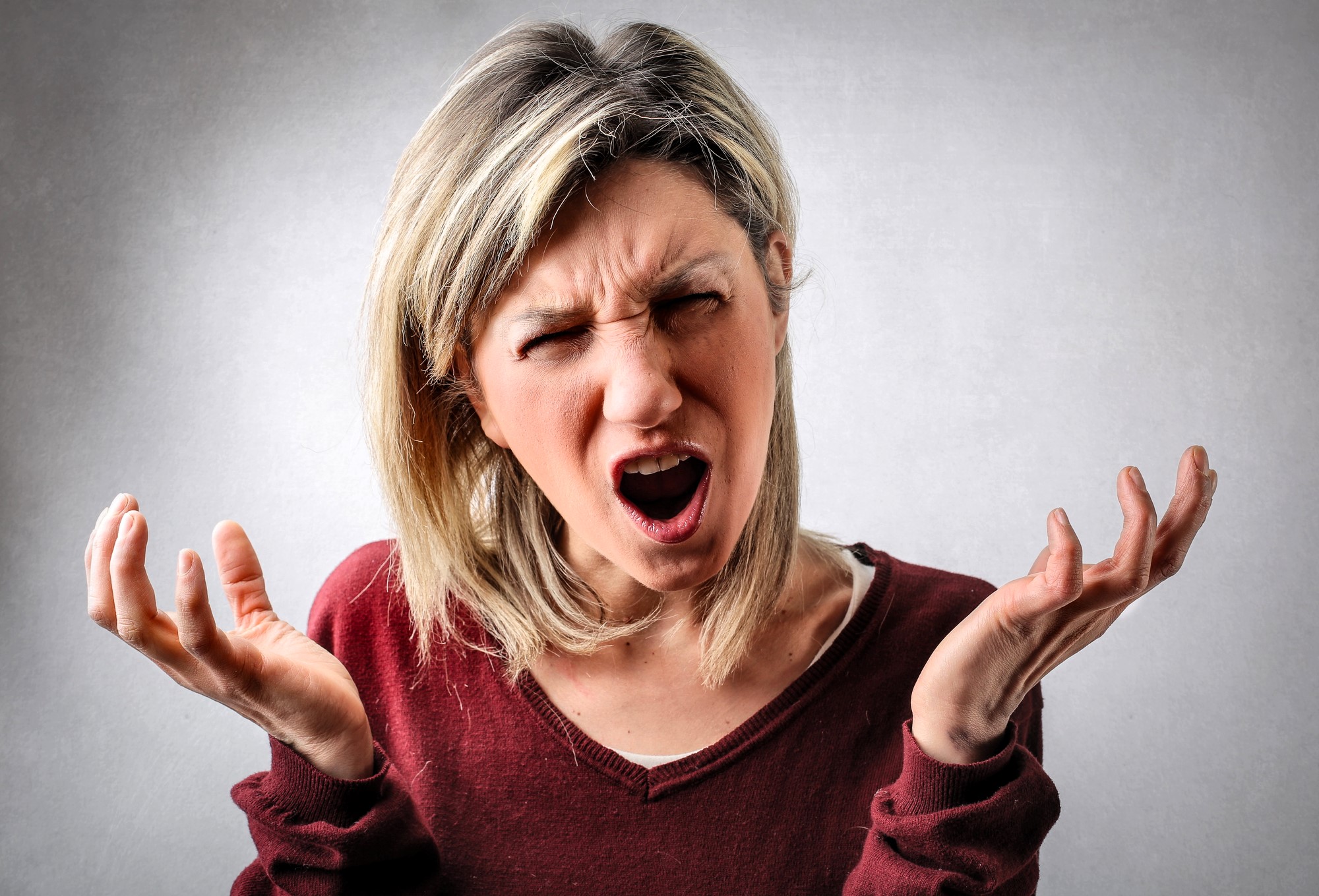 A frustrated woman with blond hair wearing a maroon top stands against a gray background. She has an open mouth and furrowed brows, raising both hands in an expressive gesture.