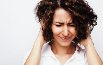 A woman with curly hair and a white shirt covers her ears with her hands, squeezing her eyes shut and frowning, as if reacting to a loud noise or discomfort. The background is plain white.