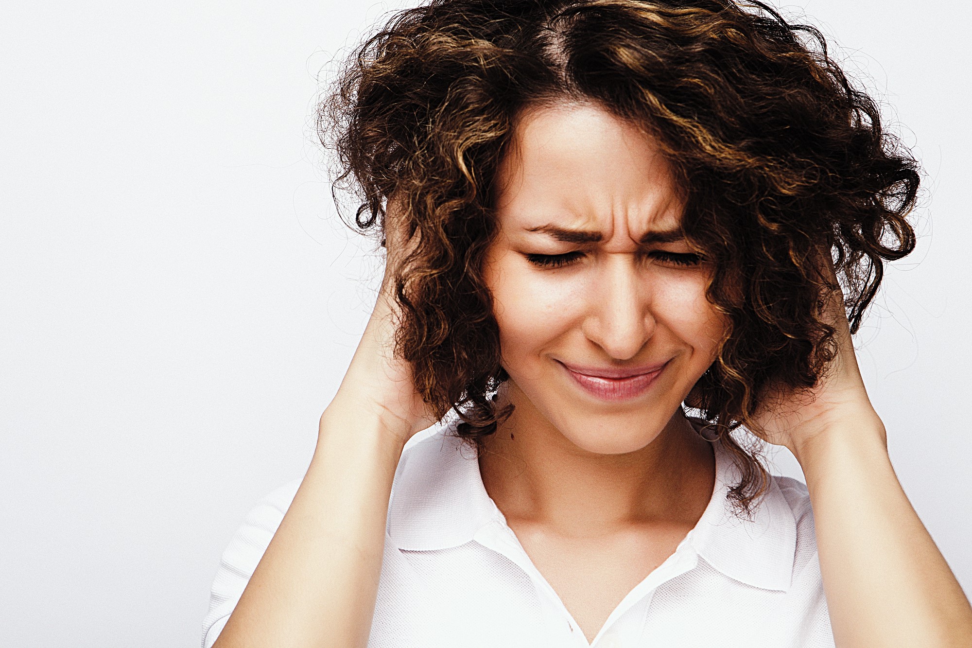 A woman with curly hair and a white shirt covers her ears with her hands, squeezing her eyes shut and frowning, as if reacting to a loud noise or discomfort. The background is plain white.