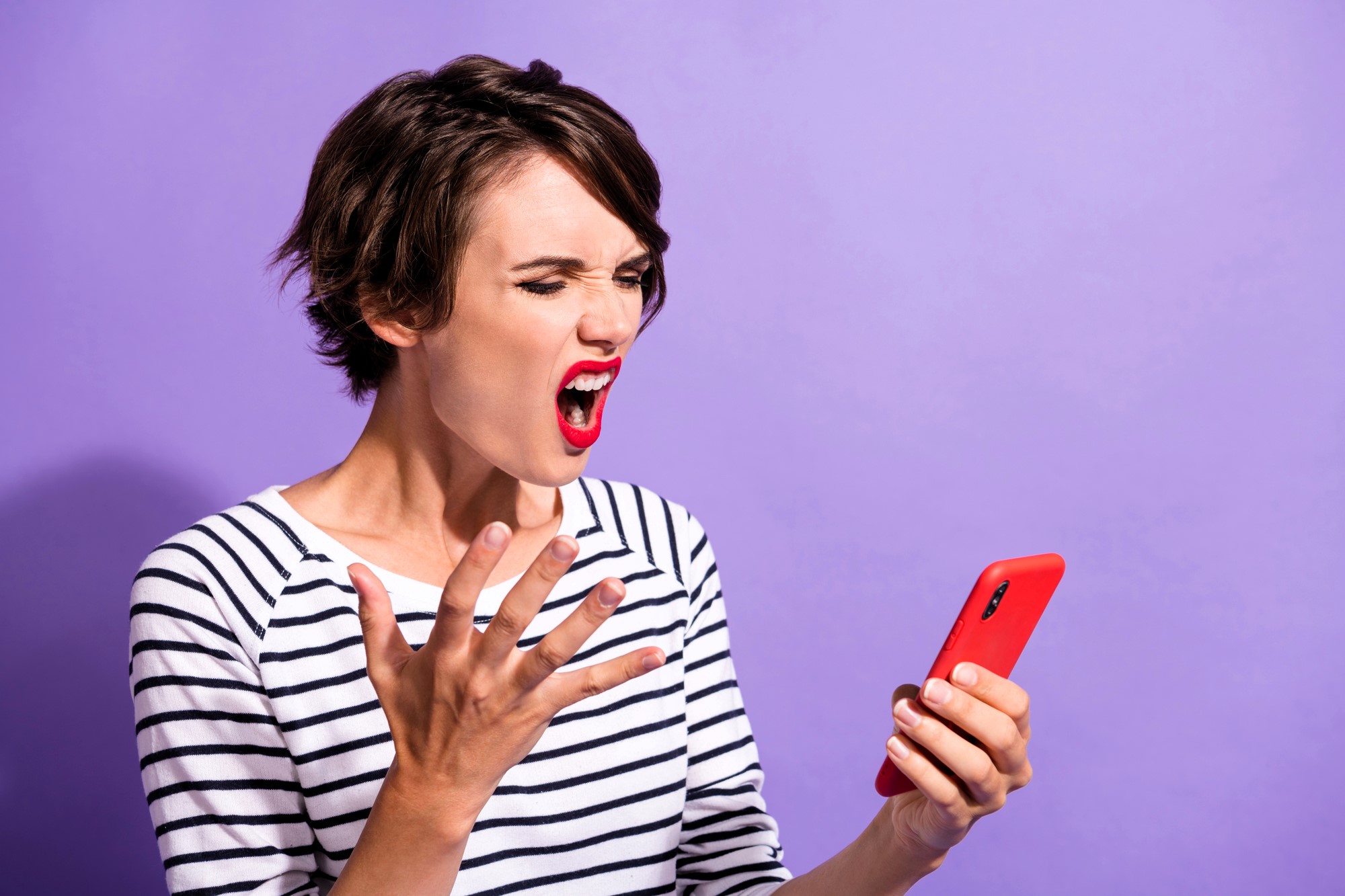 A woman with short brown hair is wearing a striped shirt and red lipstick. She looks frustrated, yelling while holding a red smartphone in one hand. Her other hand is raised with fingers splayed against a purple background.