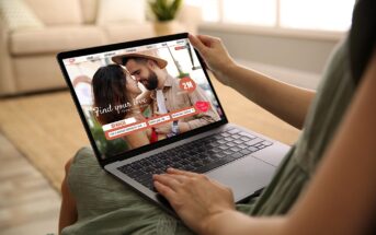 Person holding a laptop displaying an online dating website. The screen shows a couple close together, with text reading "Find your love" and "Search." The background is a cozy living room with a sofa and blurred furnishings.