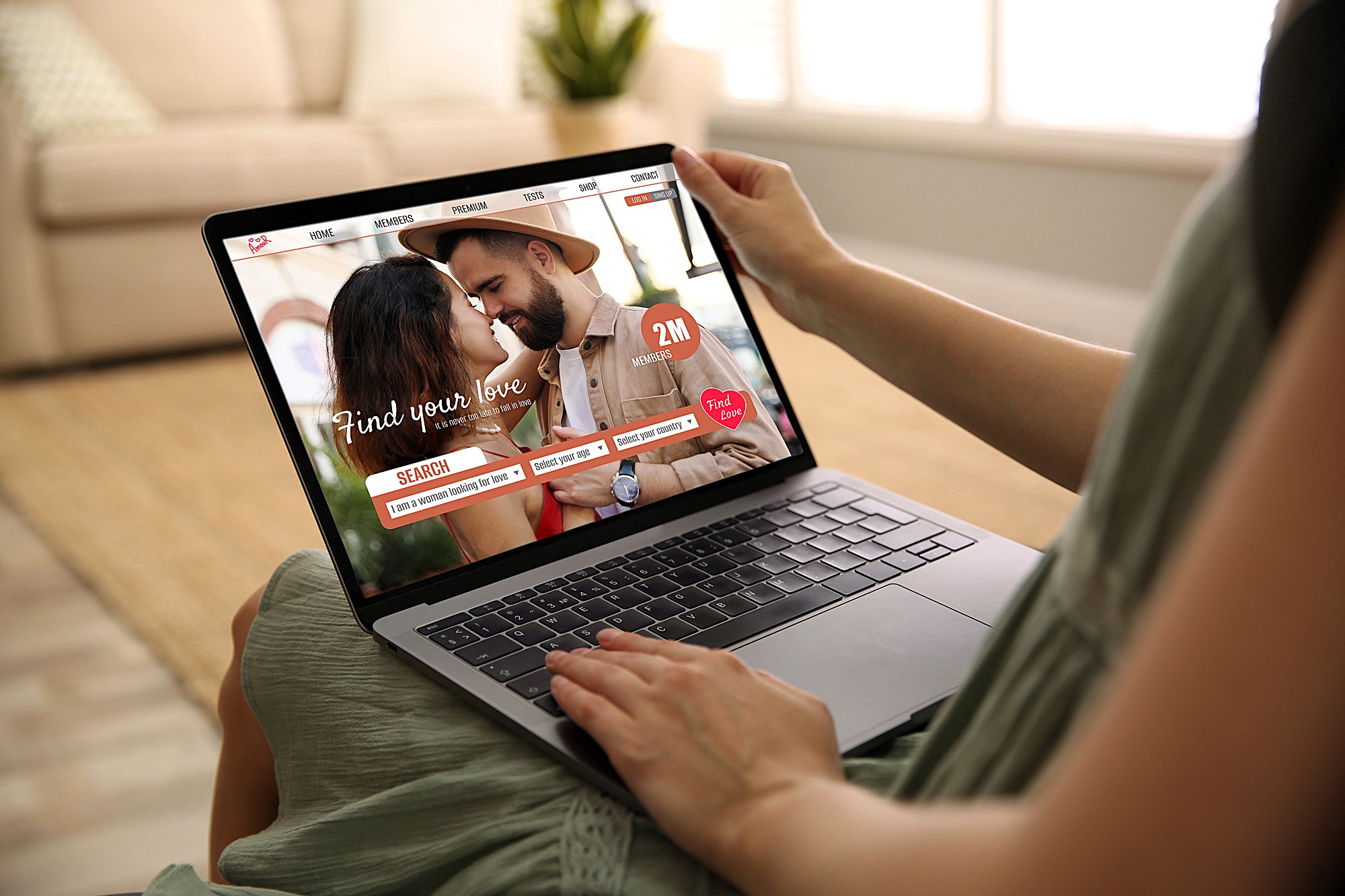 Person holding a laptop displaying an online dating website. The screen shows a couple close together, with text reading "Find your love" and "Search." The background is a cozy living room with a sofa and blurred furnishings.