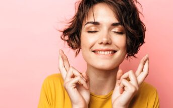 A person with short, wavy hair smiles with closed eyes and crosses their fingers, wearing a yellow shirt. The background is a light pink color.