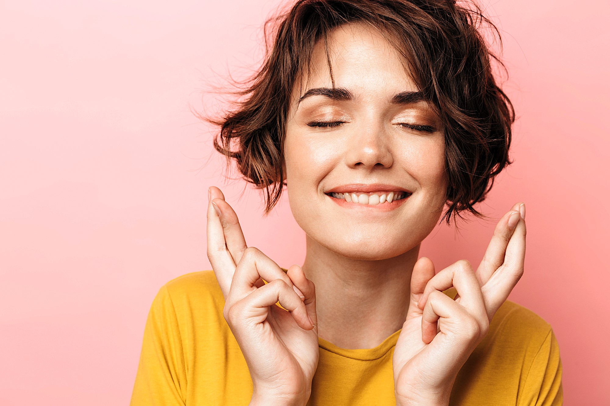 A person with short, wavy hair smiles with closed eyes and crosses their fingers, wearing a yellow shirt. The background is a light pink color.