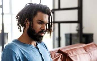 A man with dreadlocks and a beard wearing a blue shirt is sitting on a sofa, looking thoughtful. The room has large windows and natural light.