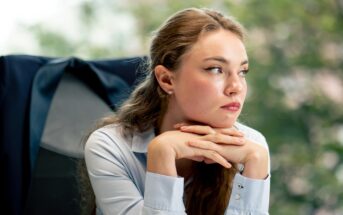 A woman with long brown hair sits at a desk, wearing a light blue blouse. She leans her chin on her hands and gazes thoughtfully to the side. A dark jacket is draped over the chair, and a blurred background of greenery is visible.